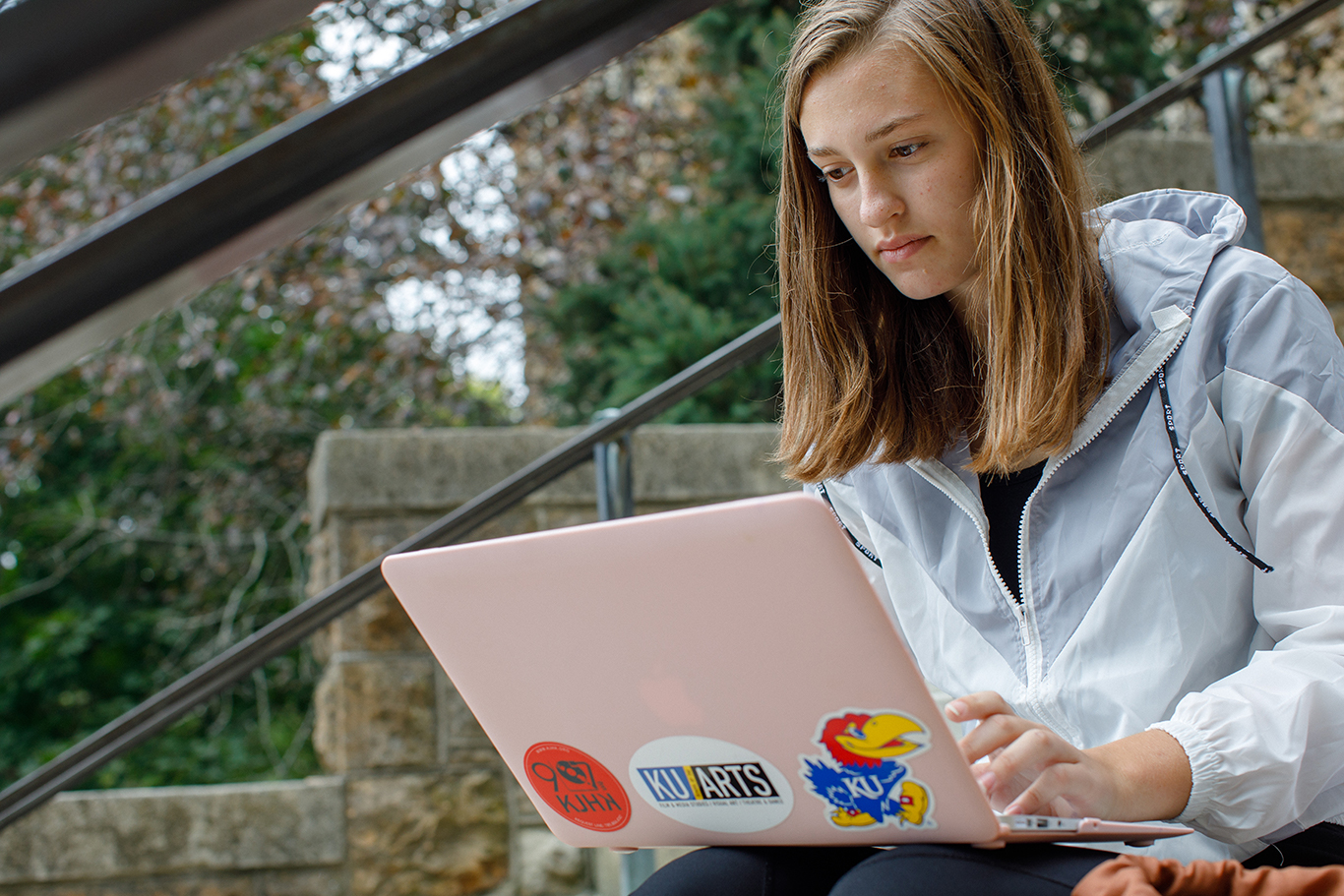 A student uses their laptop while sitting on the steps of a building. The laptop has a pink cover and multiple KU stickers on it. 