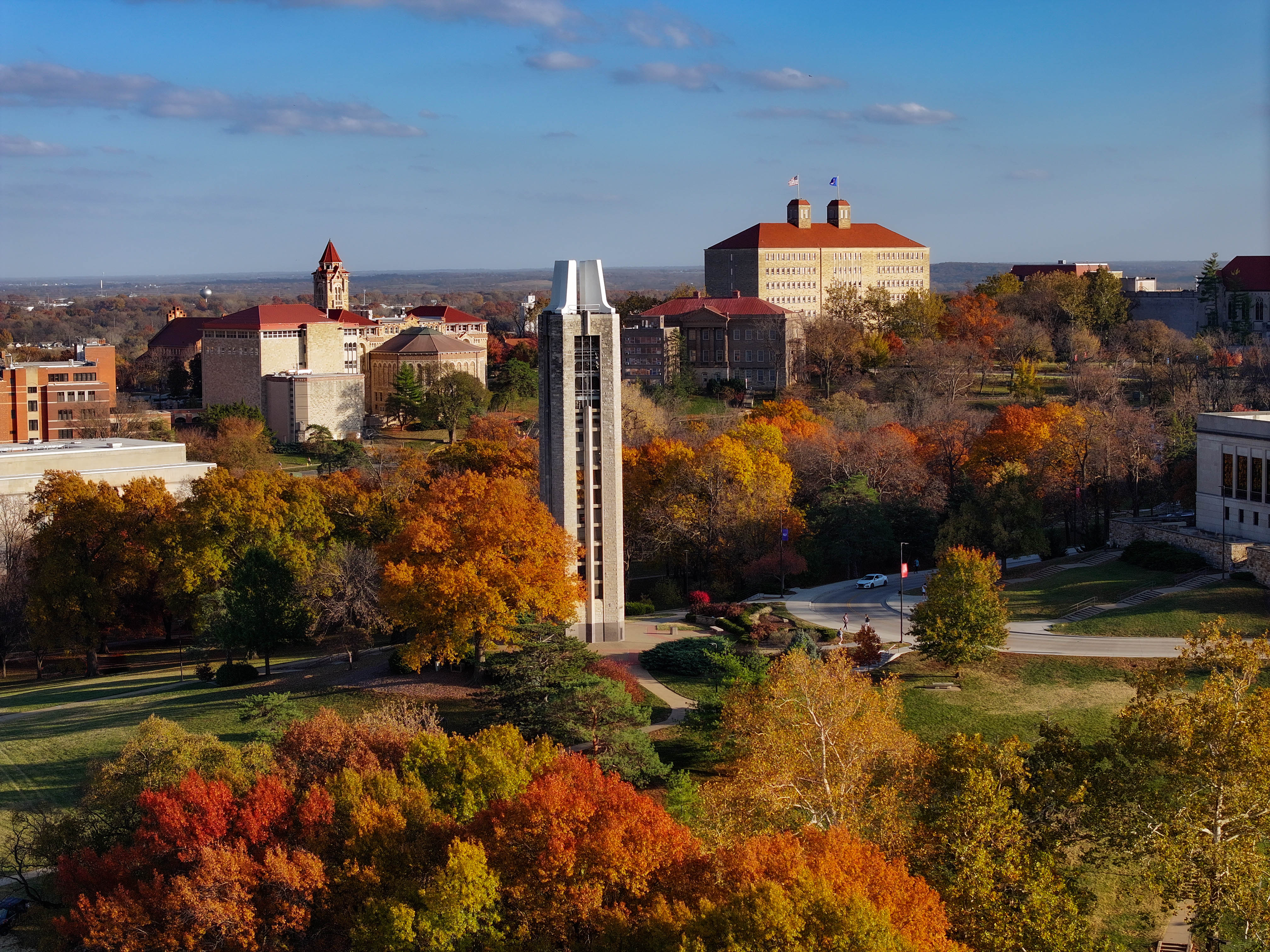 Aerial view of the University of Kansas campus during fall, showcasing the Campanile bell tower surrounded by vibrant autumn foliage in shades of red, orange, and yellow. Historic university buildings with red rooftops are visible in the background under a clear blue sky.