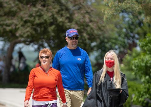 A student in a graduation cap and gown walks with their parents on a sidewalk